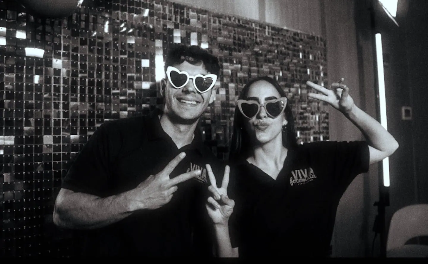 Two guests with heart sunglasses in front of a sequin backdrop, in black and white