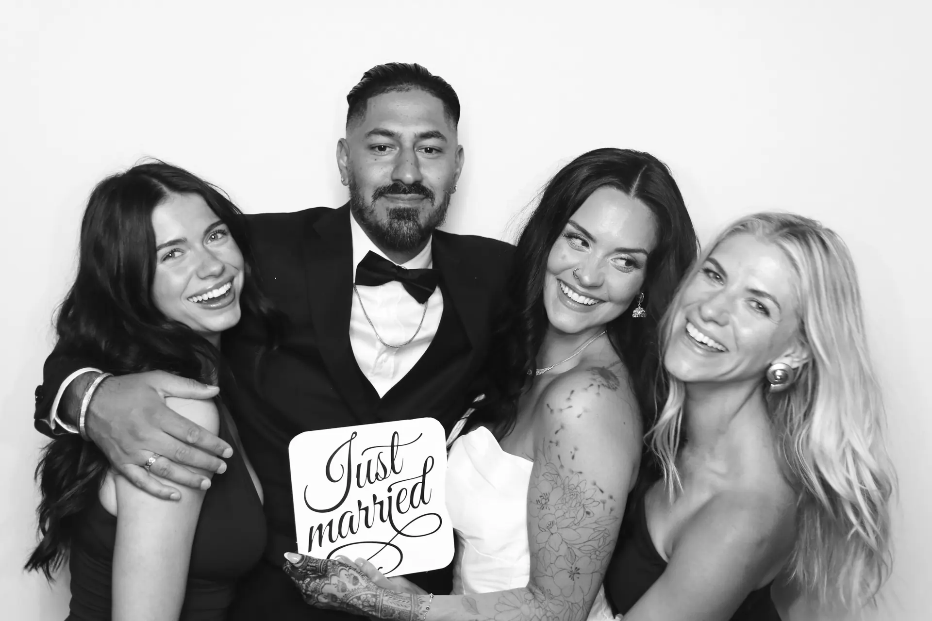 A groom in a tuxedo and three guests holding a "Just married" sign at the Viva photo booth, in black and white