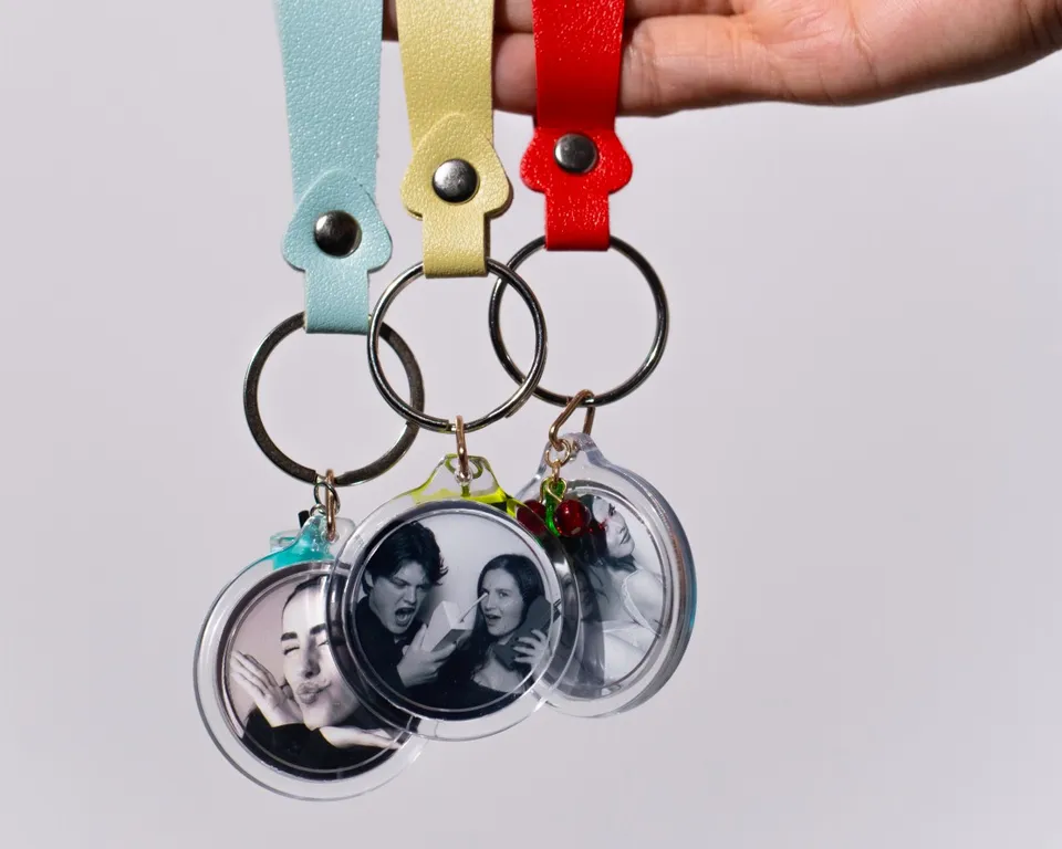 A hand holding three photo keychains on mint, yellow and red leather straps, each with a guest's black-and-white booth photo inside a clear round case