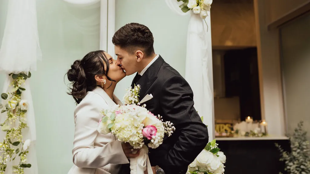 Newlyweds kissing under a white floral arch, the bride holding a white and pink bouquet